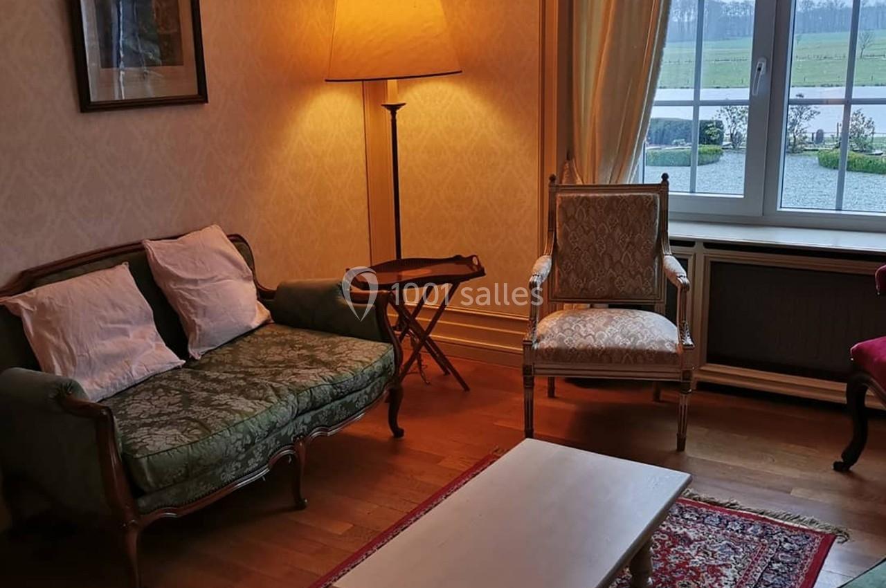 Salon classique avec canapé, fauteuil, table basse, lampe allumée et vue sur un jardin par une fenêtre.