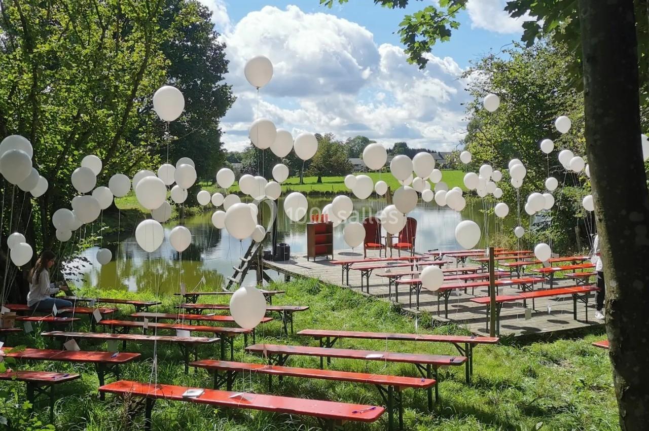 Bancs rouges alignés près d'un étang, décorés de ballons blancs flottants sous un ciel partiellement nuageux.
