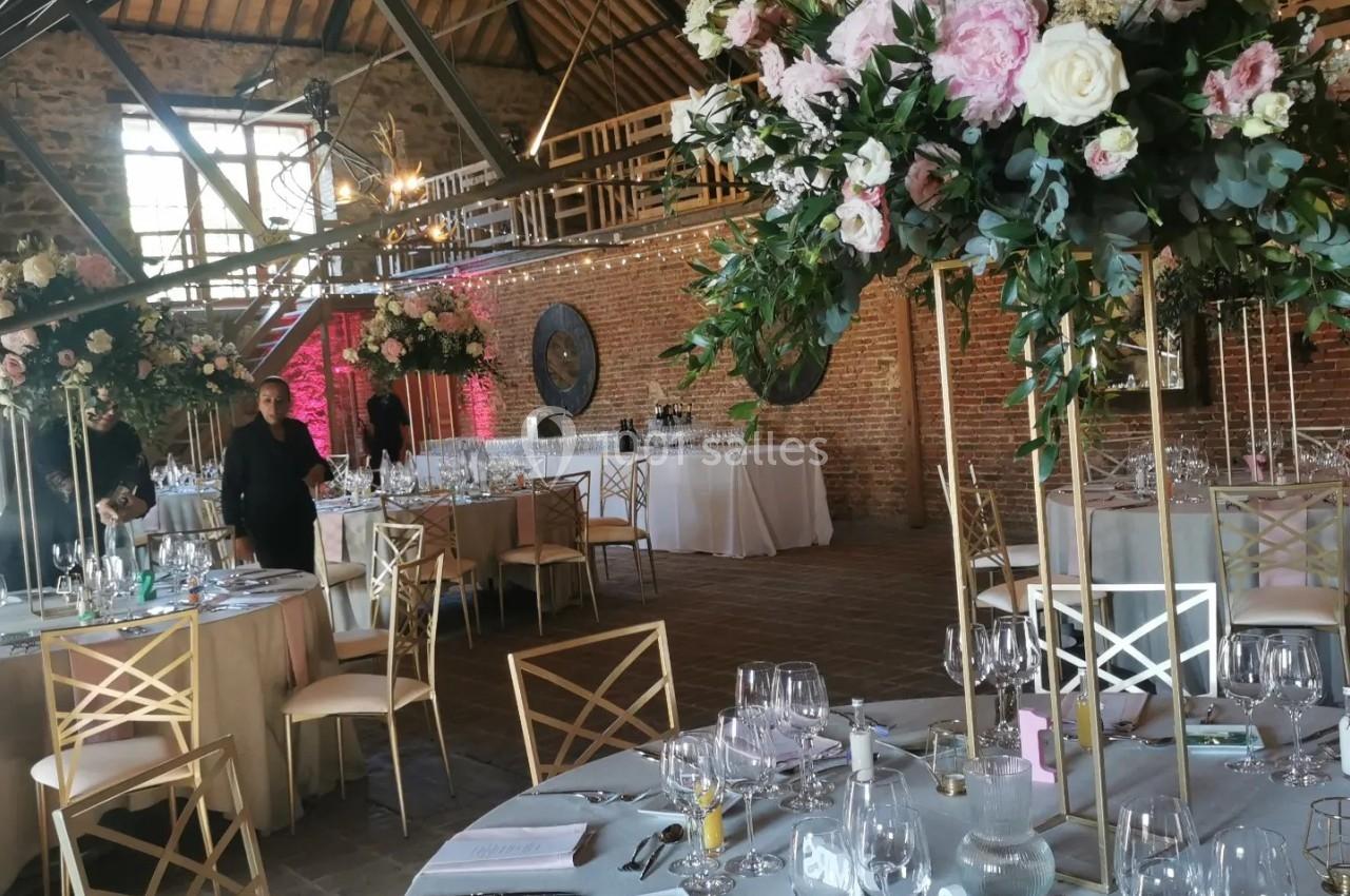 Salle de réception décorée pour un mariage, avec tables dressées, fleurs et éclairage naturel sous une charpente en bois.