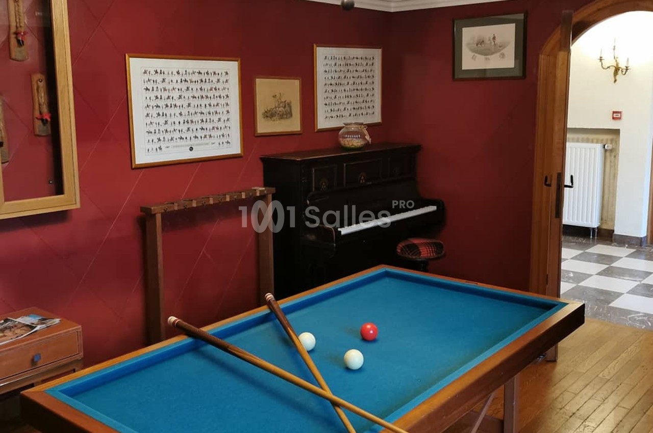 Table de billard avec trois boules et deux queues, dans une pièce avec un piano, des cadres et des murs rouges.