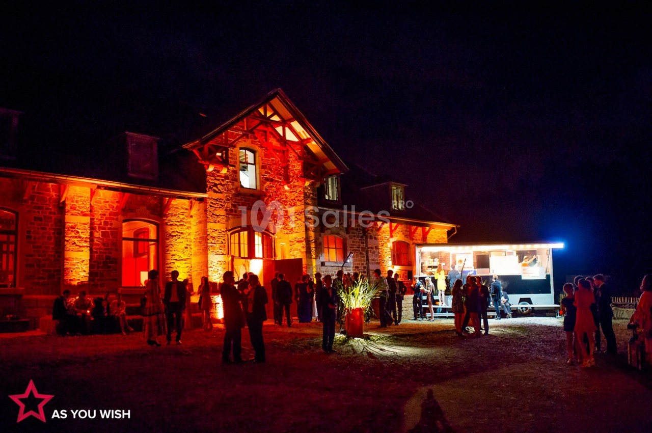 Façade d'un bâtiment en pierre éclairée en rouge, avec un food truck et des groupes de personnes rassemblées la nuit.