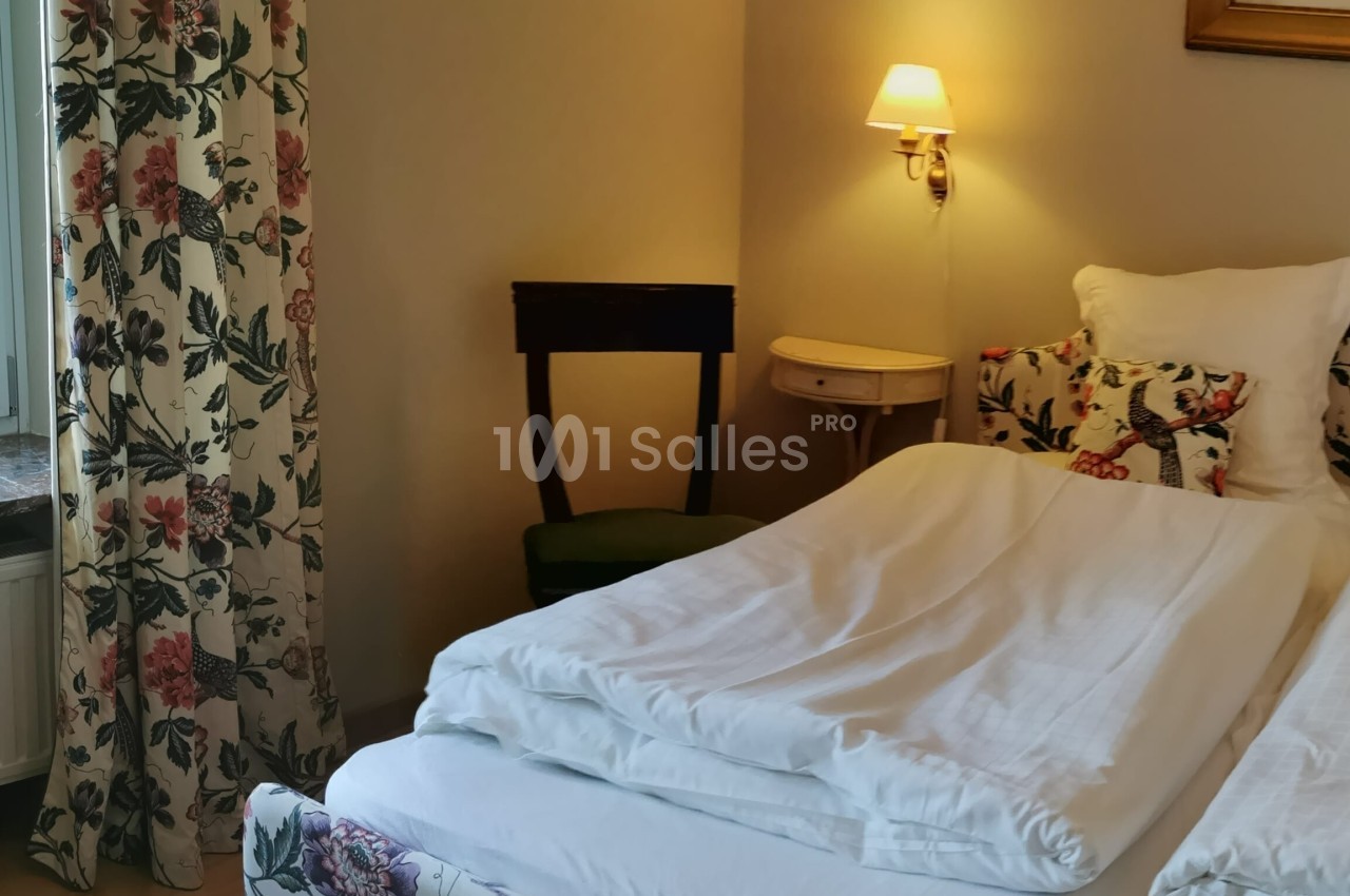 Lit double avec draps blancs dans une chambre décorée de motifs floraux, éclairée par une lampe murale.