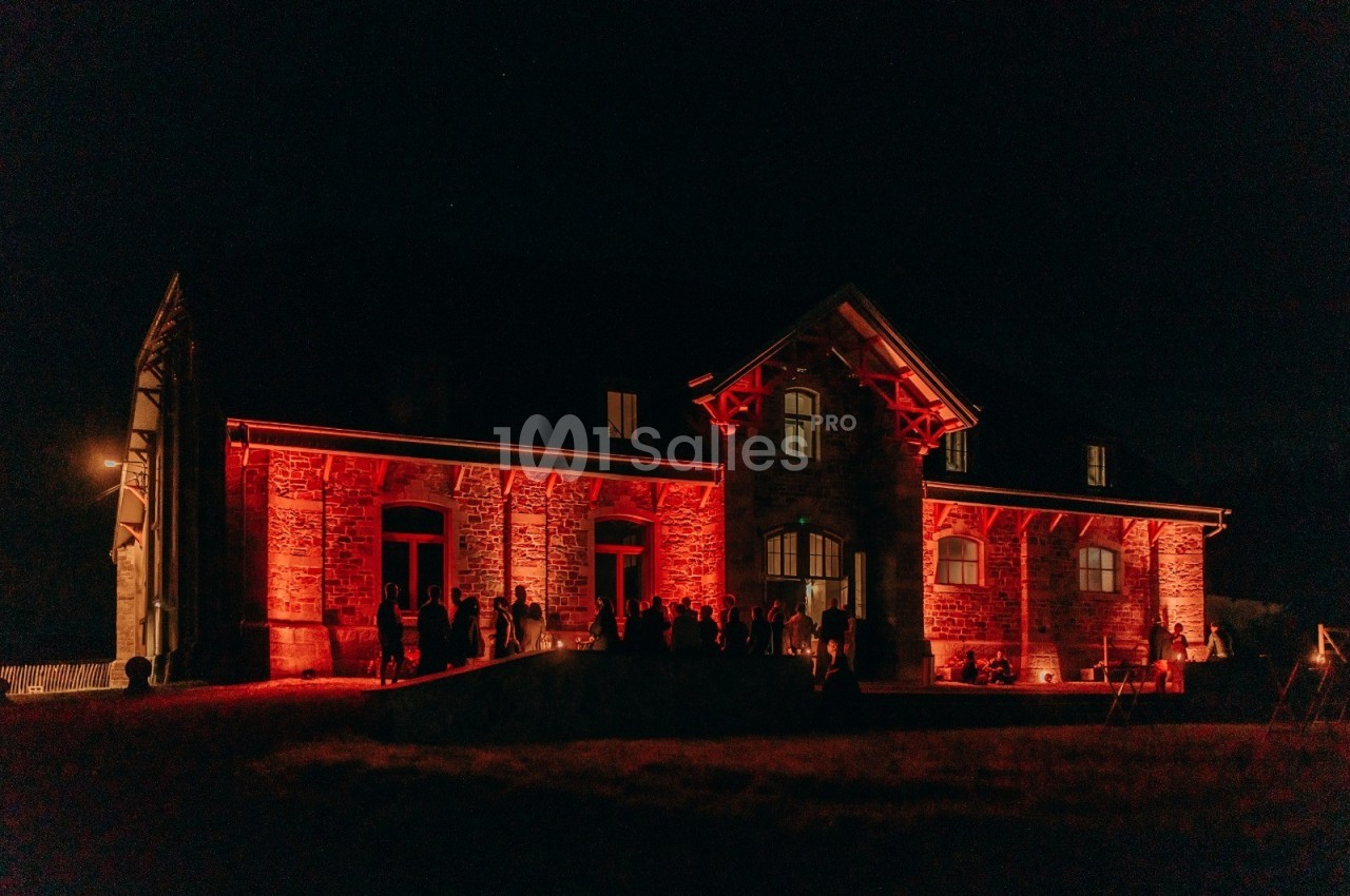 Façade d'une grande maison en pierre éclairée par des lumières rouges, avec des silhouettes de personnes devant.