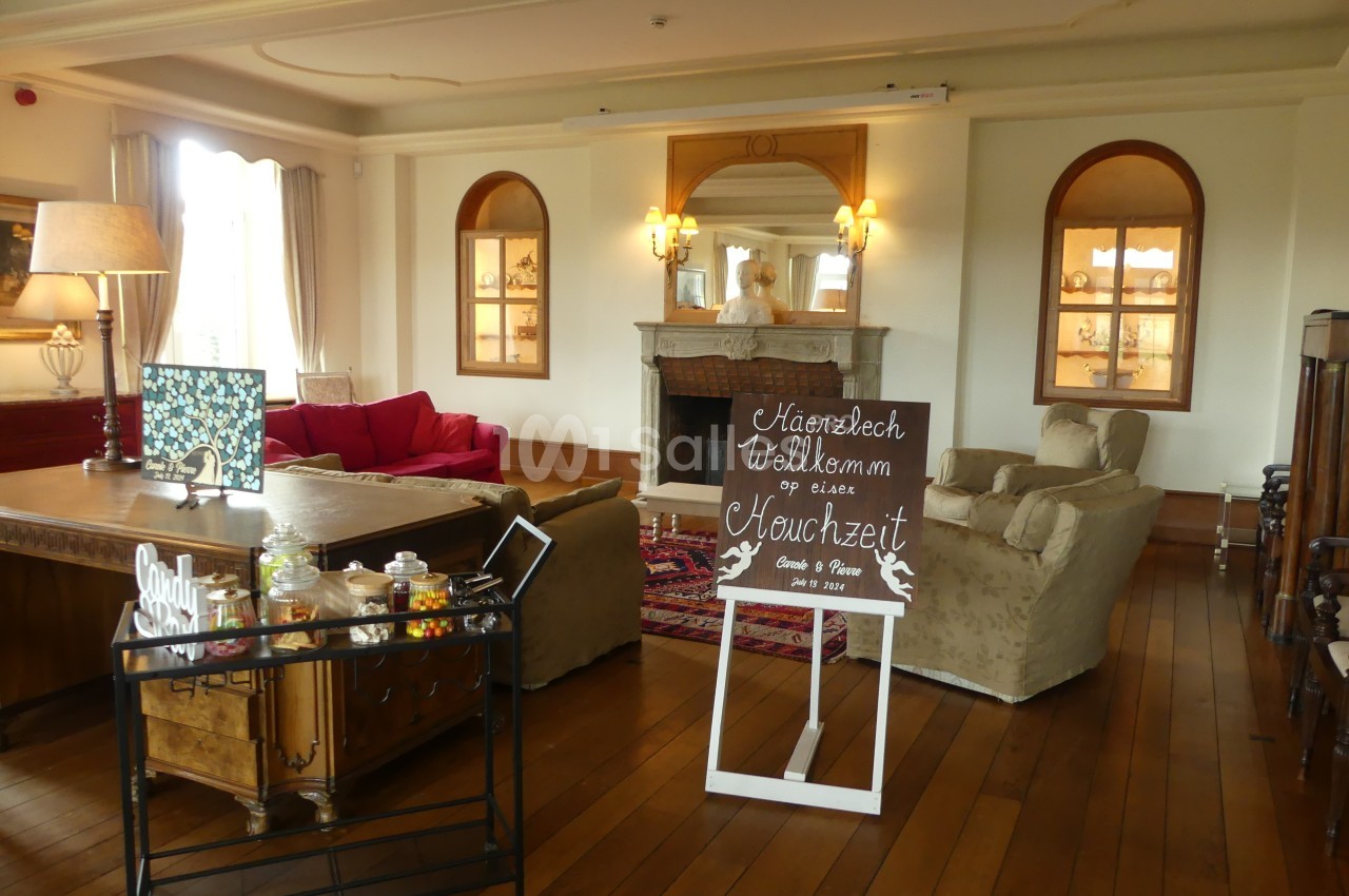 Salon élégant avec parquet, cheminée, canapé rouge, fauteuils beiges et panneau de bienvenue pour un mariage.