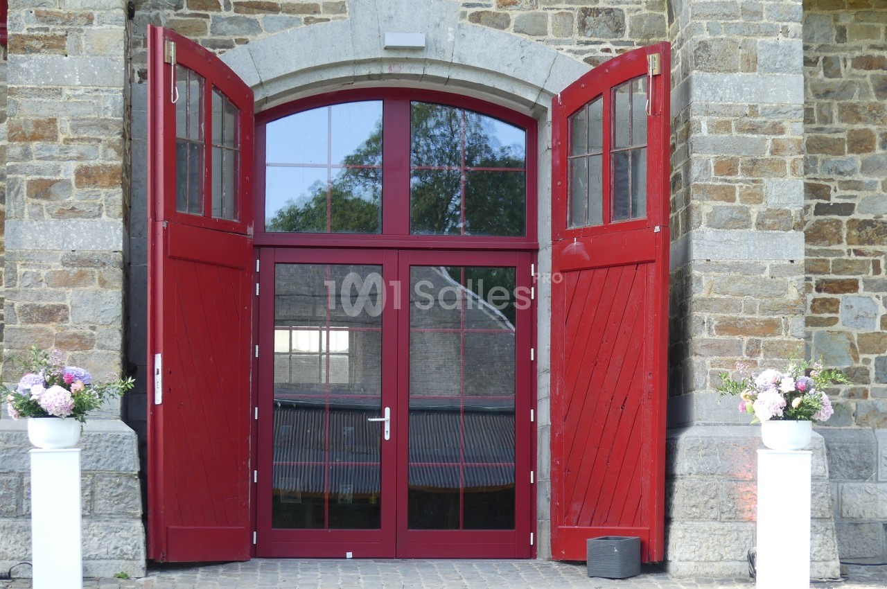 Porte d'entrée rouge à double battant ouverte, entourée d'un mur en pierre, avec des pots de fleurs de chaque côté.