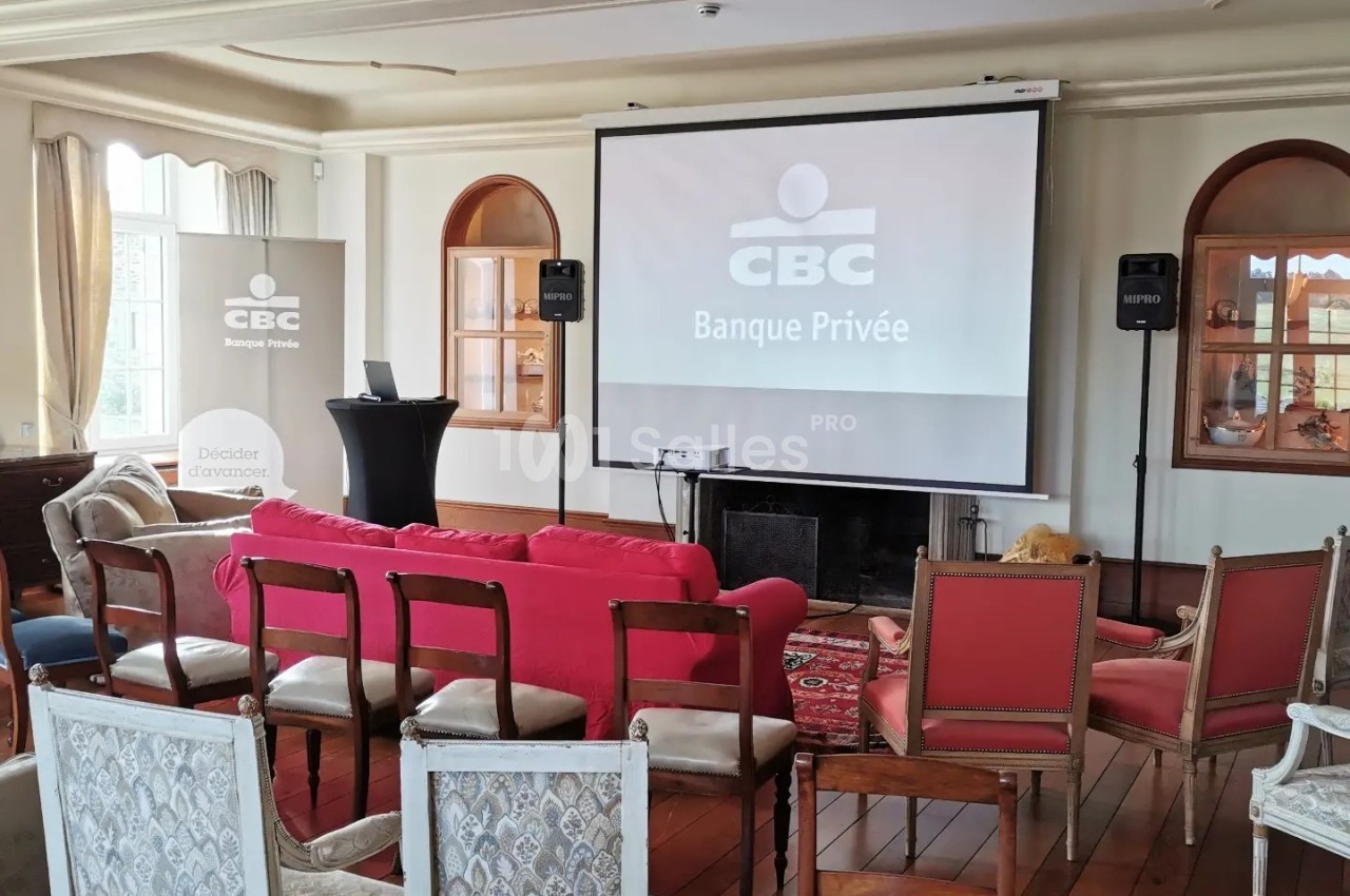 Salle de conférence avec écran de projection affichant ’CBC Banque Privée’, rangées de chaises et canapé rouge.