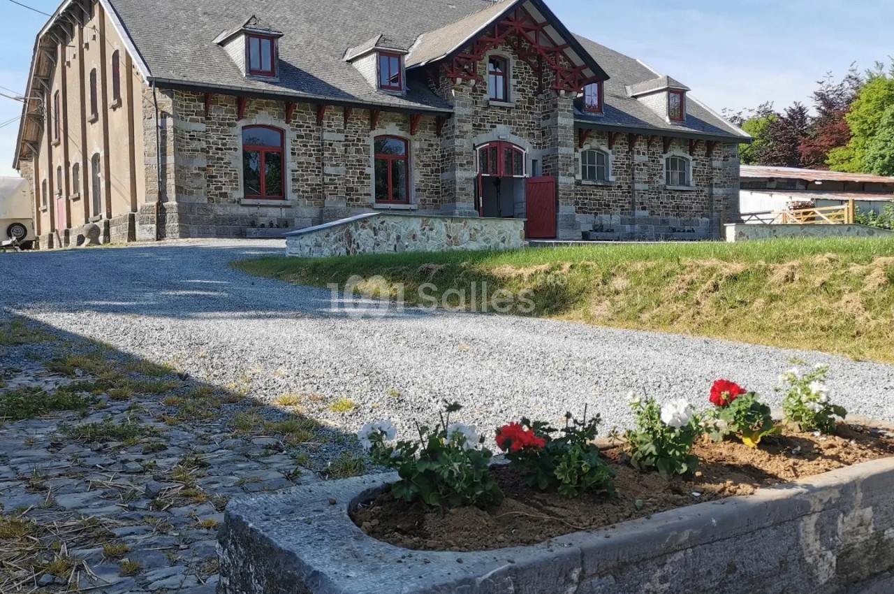 Grande maison en pierre avec toit en ardoise, allée gravillonnée et jardinière en bois contenant des fleurs rouges.