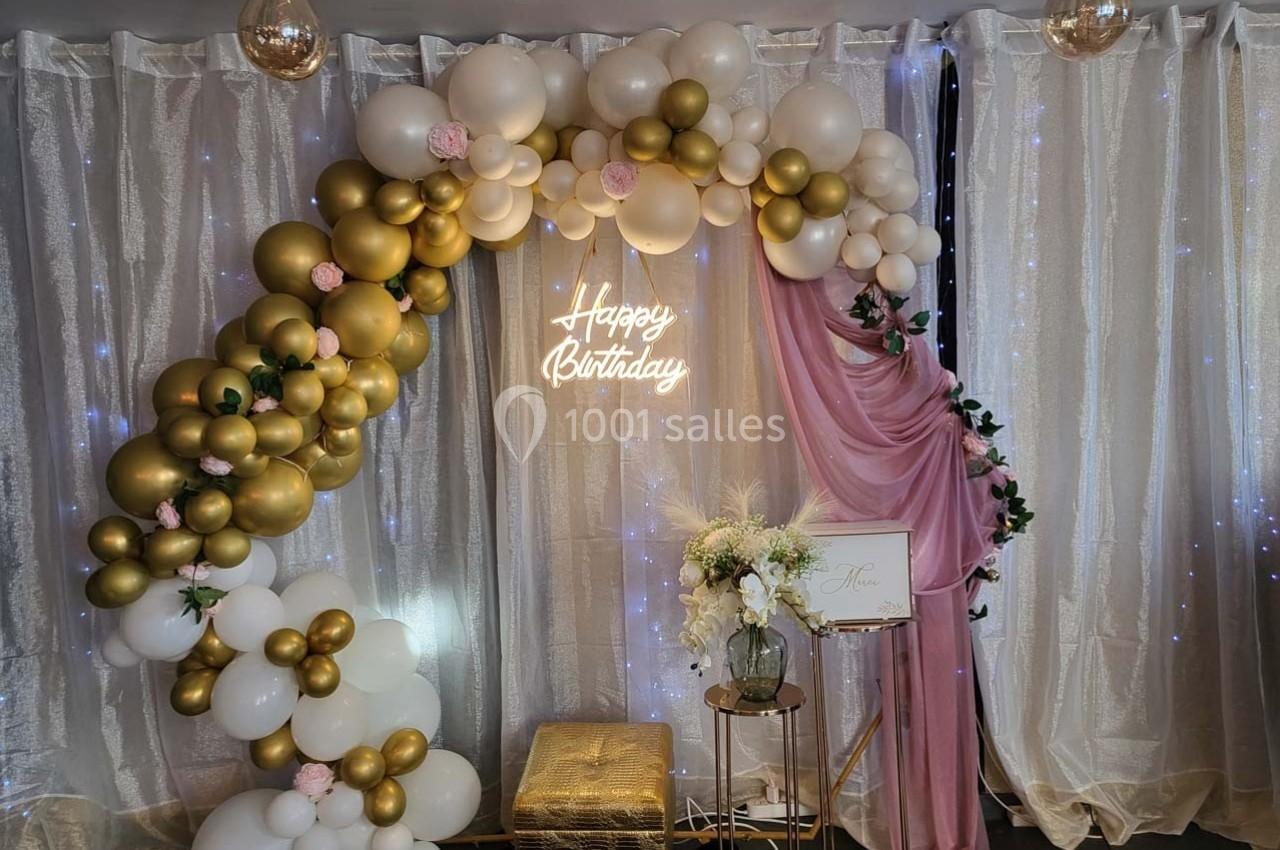 Arc de ballons blancs et dorés avec un panneau ’Happy Birthday’, décoré de fleurs, devant un rideau lumineux.