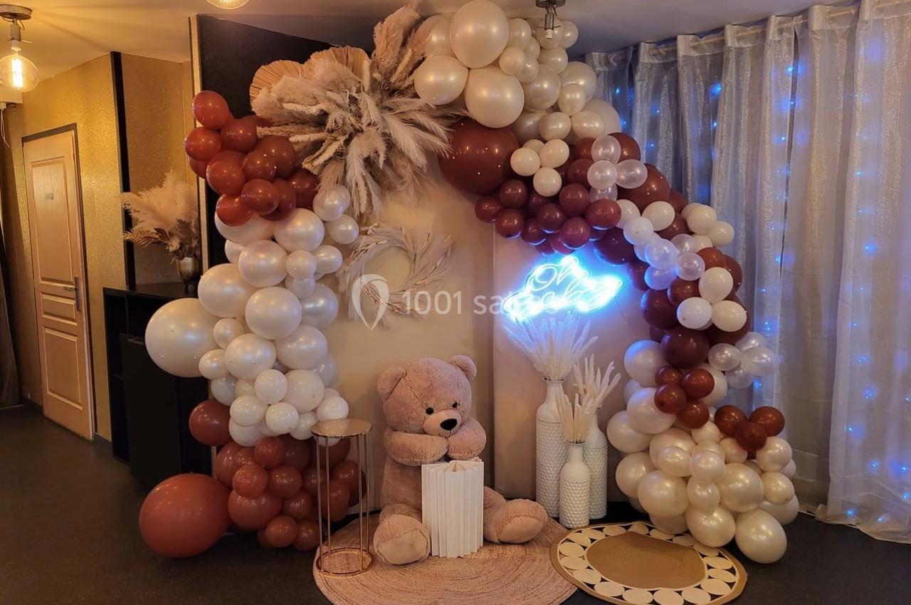 Décoration festive avec arche de ballons blancs et marron, ours en peluche, néon lumineux et éléments décoratifs.