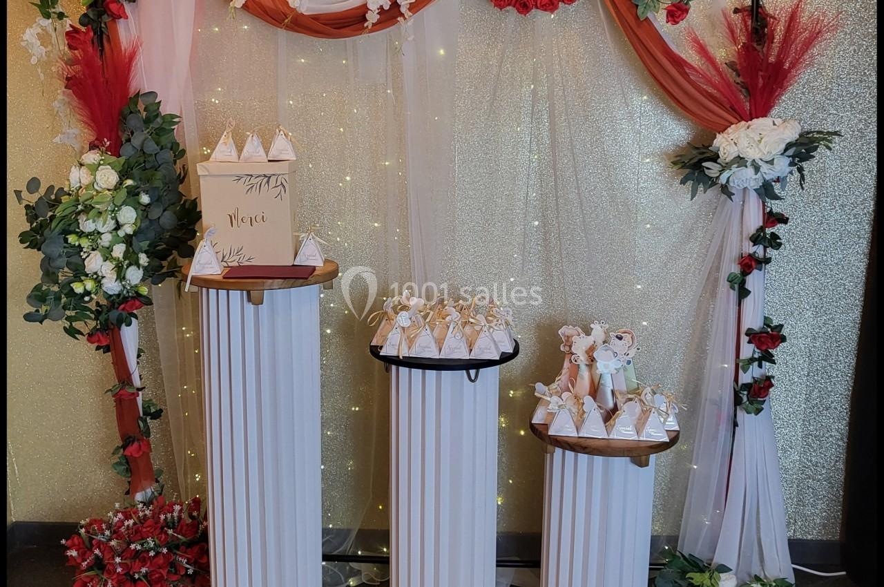 Décoration florale avec arche ornée de roses rouges et blanches, trois colonnes avec boîtes cadeaux et détails lumineux.