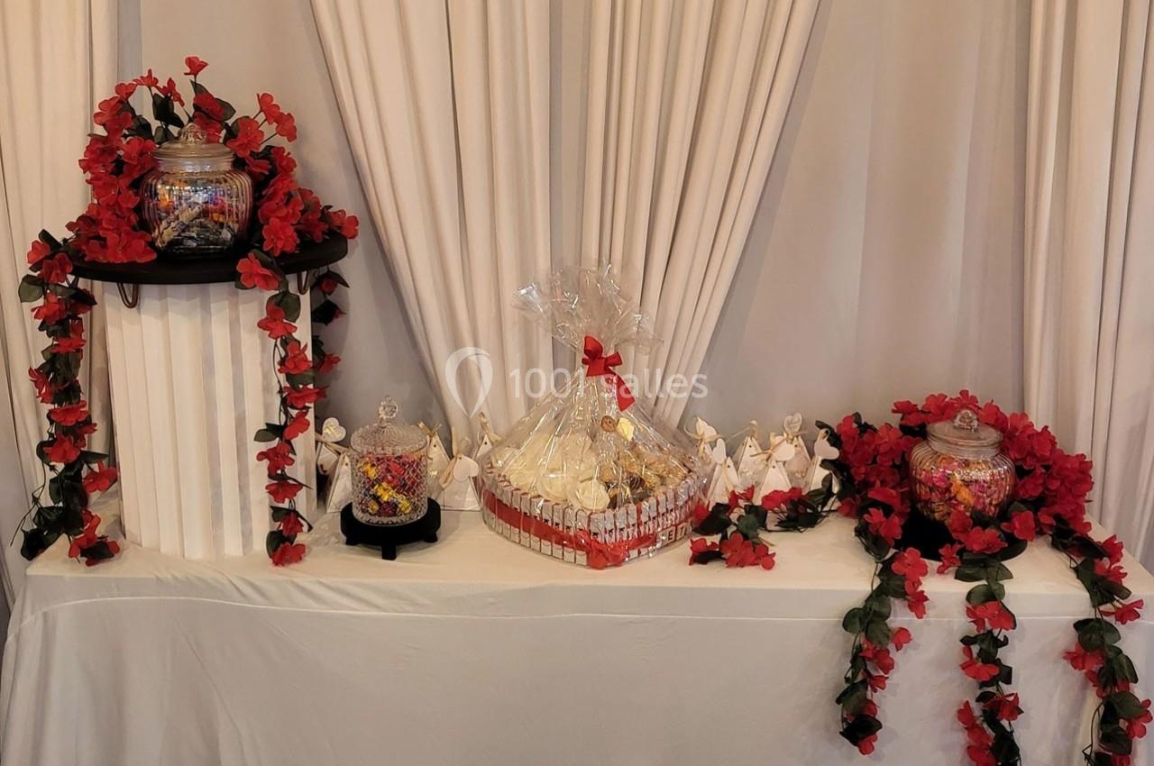 Table décorée avec des guirlandes de fleurs rouges, contenant des bonbonnières et un panier cadeau emballé.