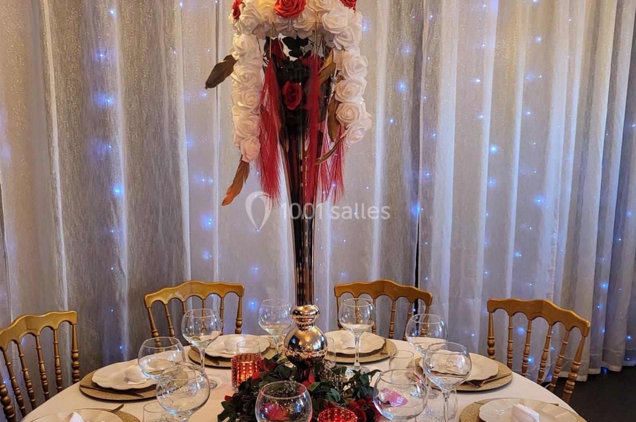 Centre de table floral avec roses rouges et blanches sur un vase haut, entouré de bougies et vaisselle sur une table dressée.