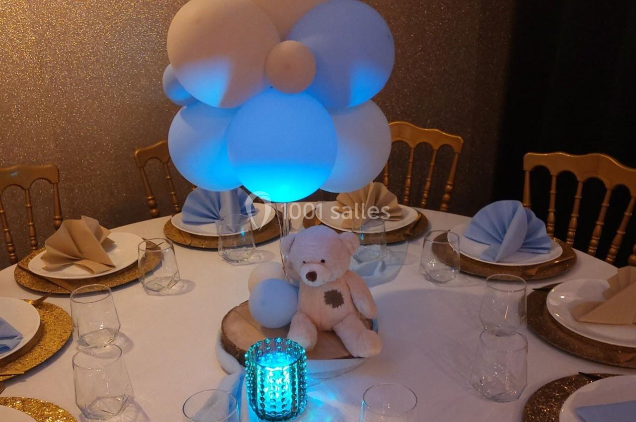 Centre de table avec ballons bleus et blancs, ourson en peluche, bougie décorative et vaisselle élégante.
