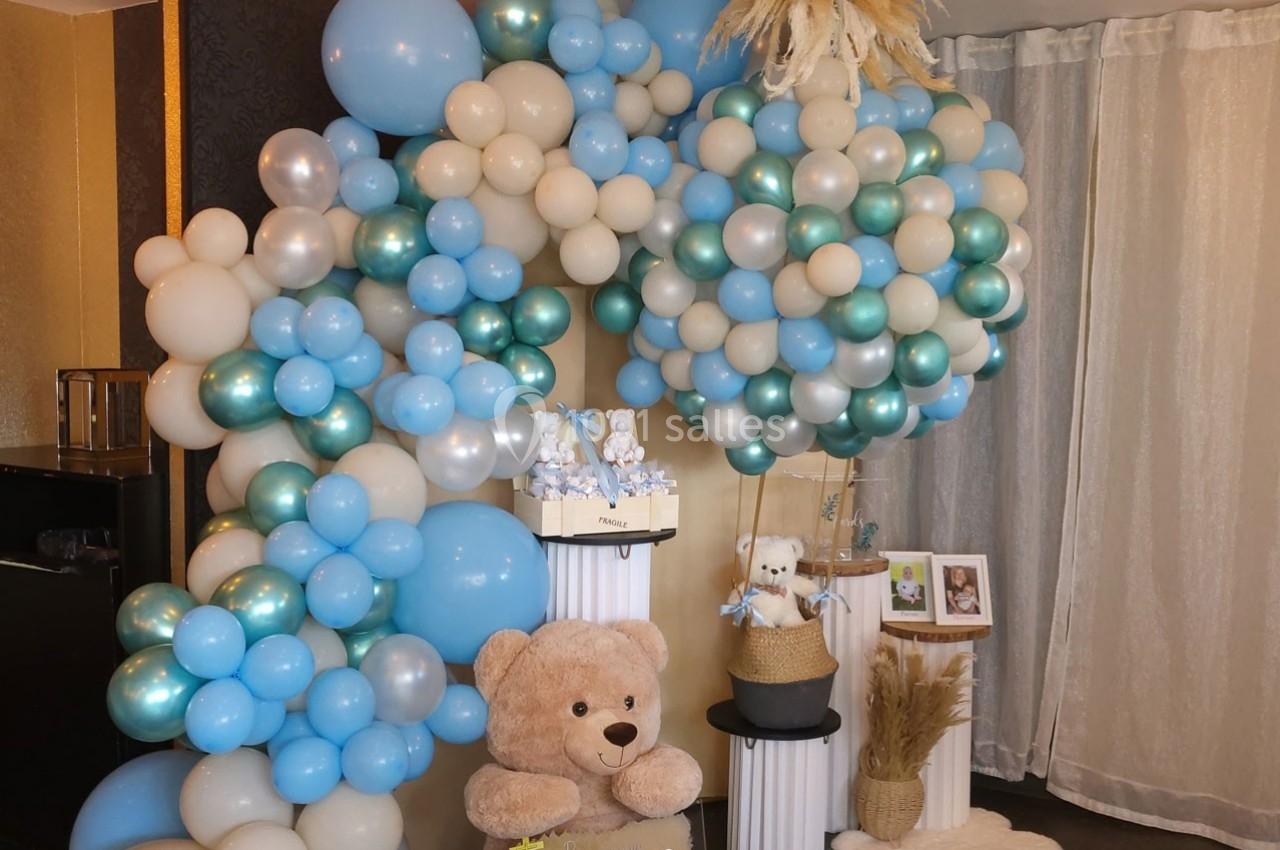 Décoration avec des ballons bleus, blancs et dorés, un ours en peluche et des éléments de fête dans une pièce lumineuse.