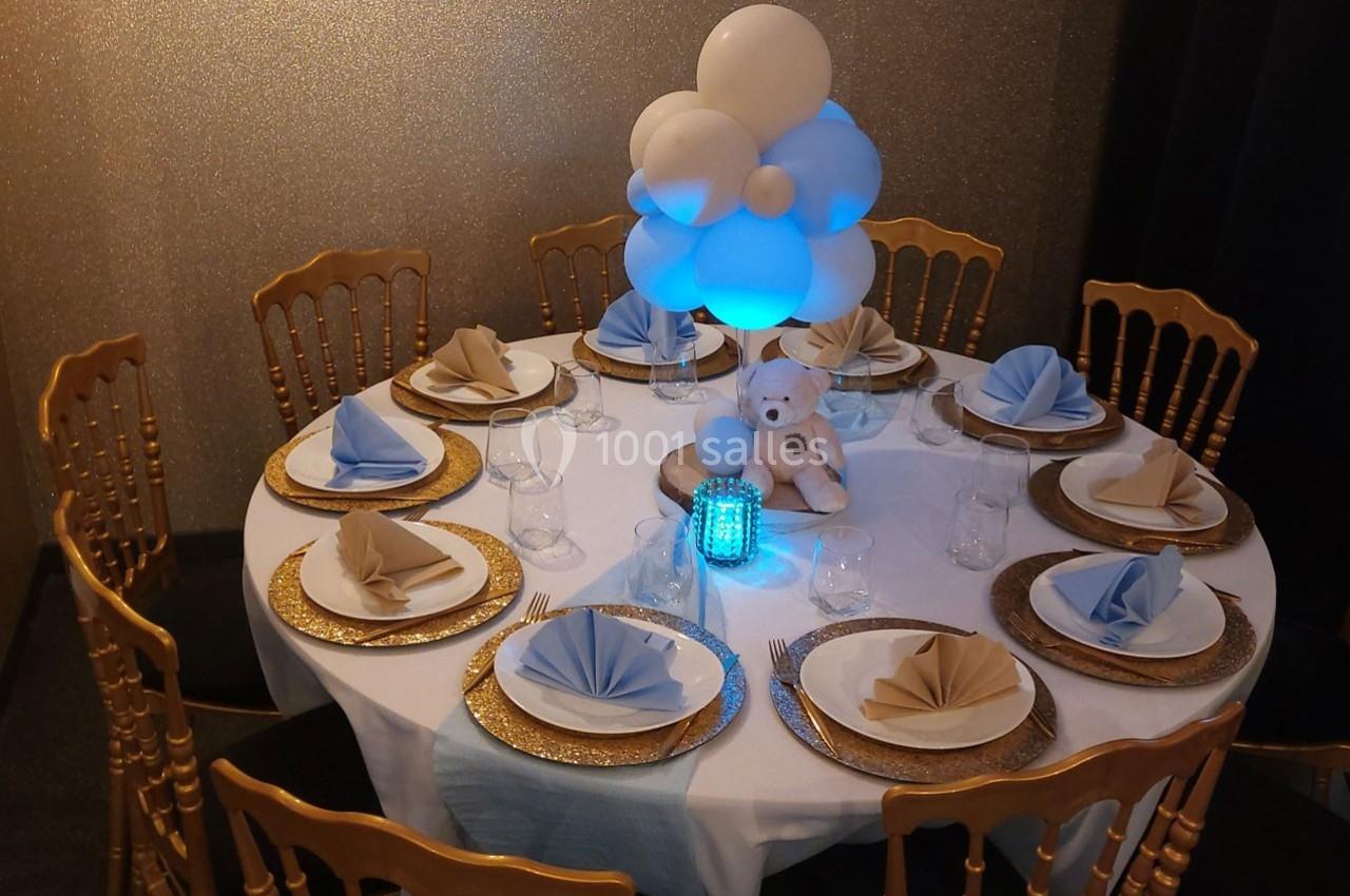 Table ronde décorée avec nappes blanches, serviettes pliées, assiettes dorées et centre de table avec ballons et peluche.