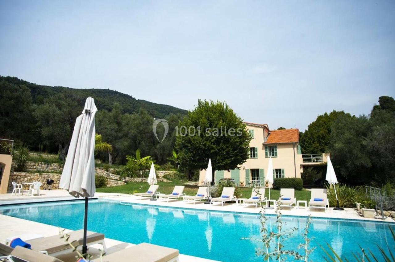 Piscine extérieure entourée de transats et parasols, avec une maison et des arbres en arrière-plan.