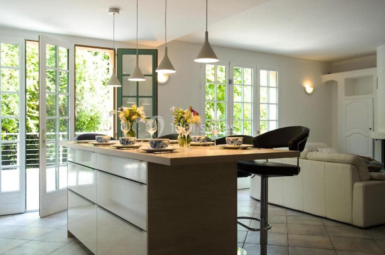 Cuisine lumineuse avec îlot central en bois, tabourets, vaisselle dressée et grandes baies vitrées donnant sur un jardin.