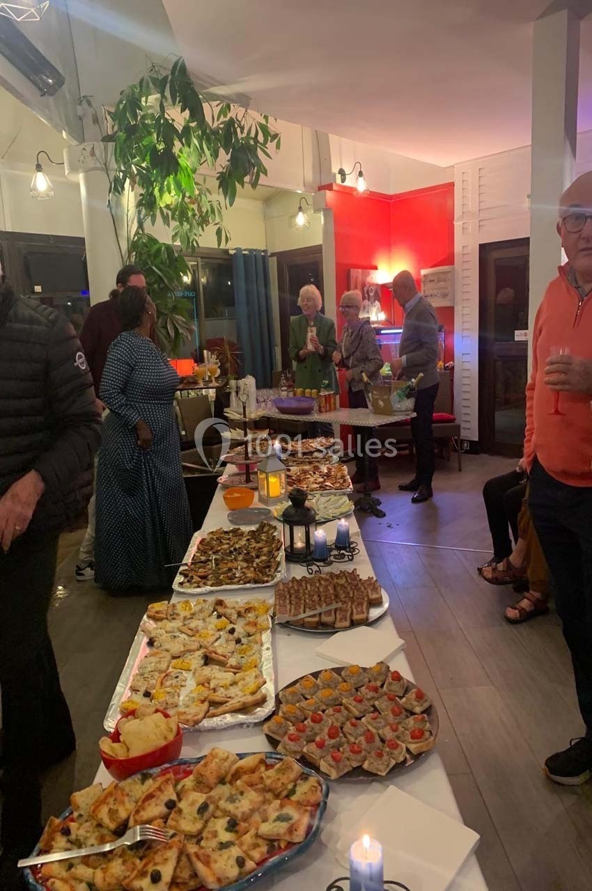 Buffet varié avec pizzas, tartines et autres plats, entouré de convives dans une salle éclairée et conviviale.