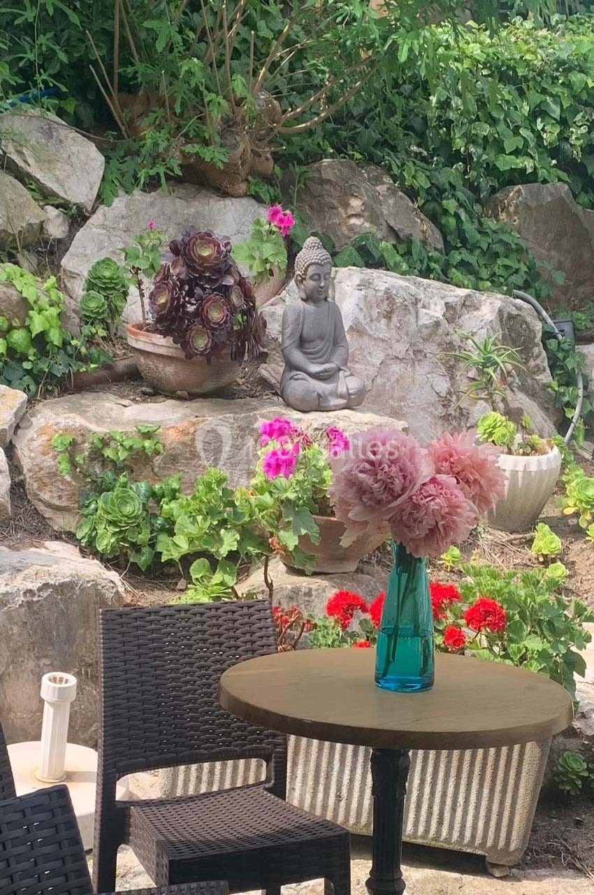 Jardin avec statue de Bouddha, fleurs colorées, table en bois et chaises en osier devant des rochers ornés de plantes.