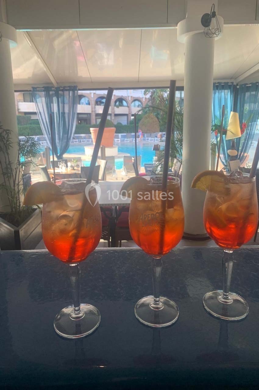 Trois verres de cocktail orange garnis de tranches d'orange et de glaçons, posés sur un bar avec vue sur une piscine.