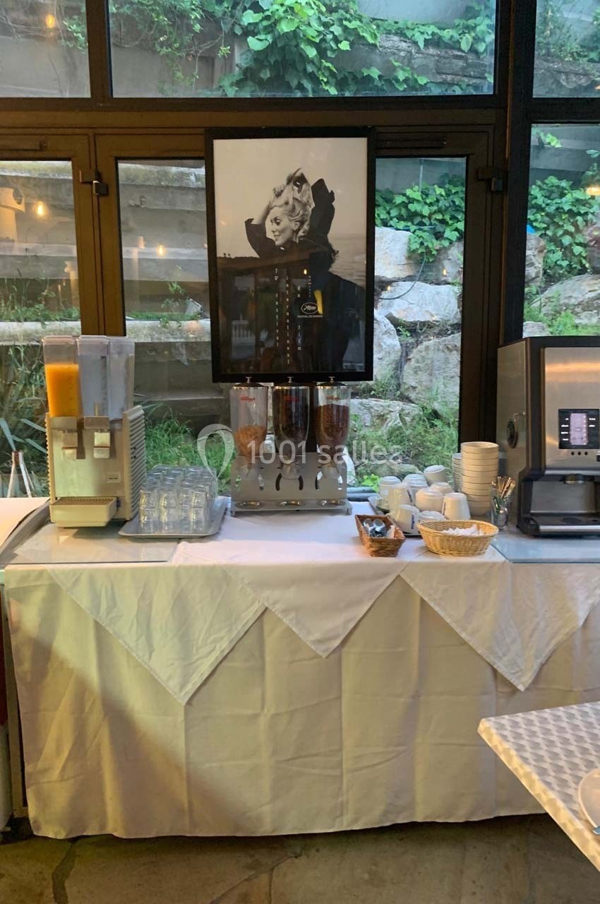Buffet de petit-déjeuner avec distributeurs de jus, céréales, machine à café et vaisselle sur une nappe blanche.