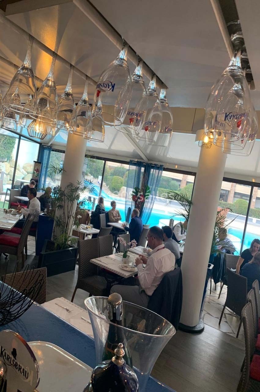 Salle de restaurant lumineuse avec vue sur une piscine, tables dressées et verres suspendus au plafond.