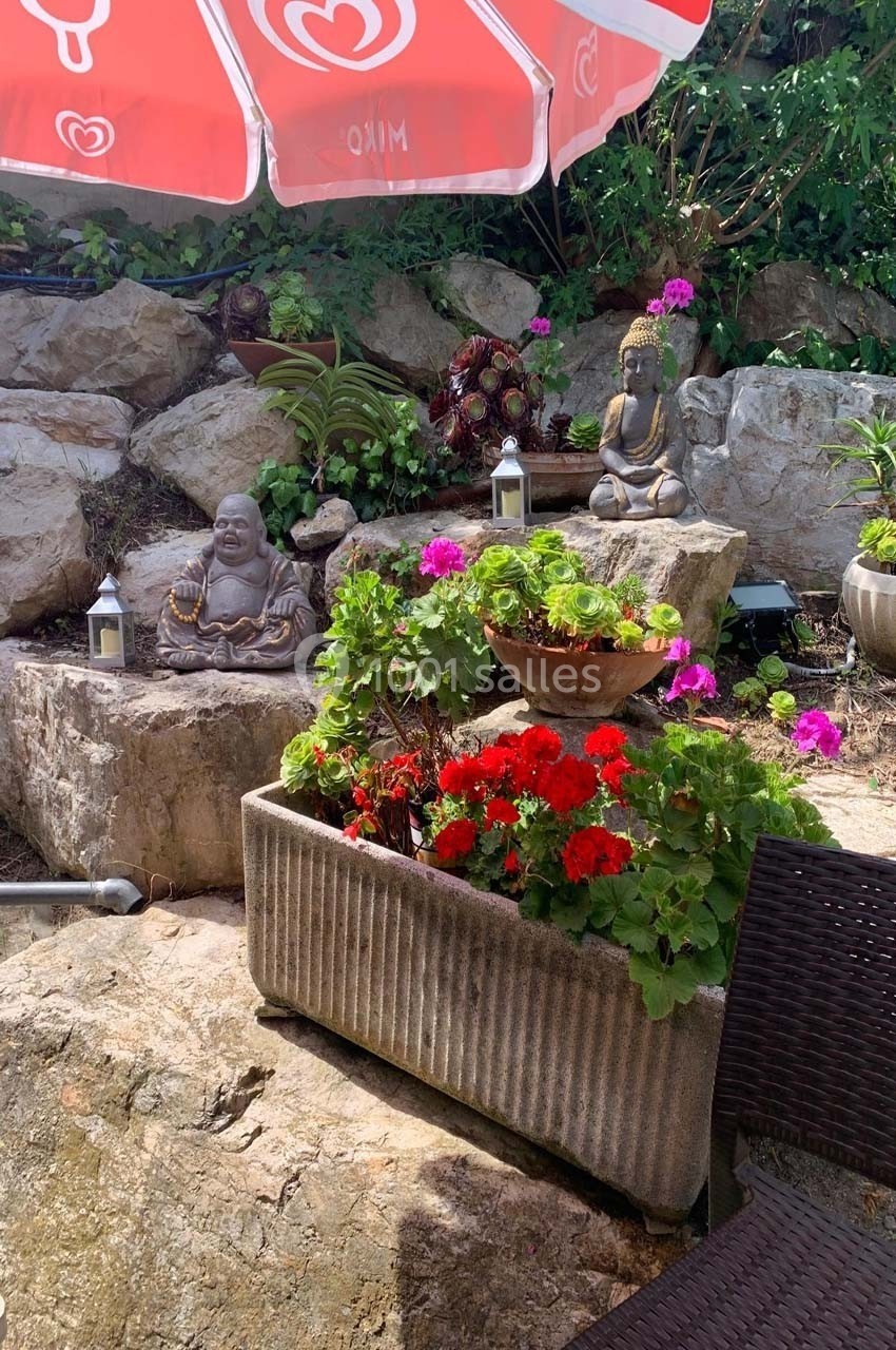 Jardin décoratif avec statues de Bouddha, fleurs colorées en jardinières et parasol rouge en arrière-plan.