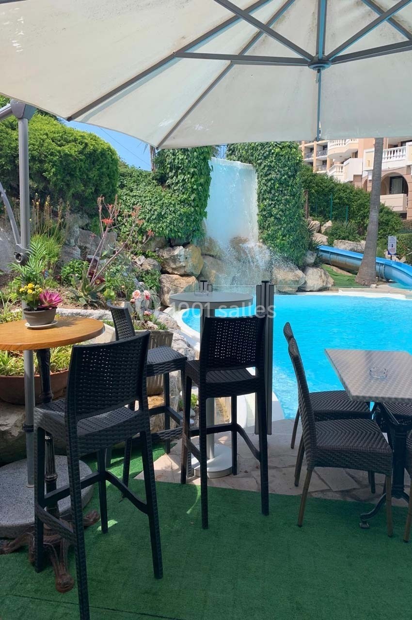 Terrasse extérieure avec tables et chaises hautes près d'une piscine et d'une cascade entourée de végétation.