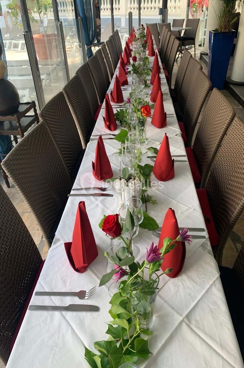 Table dressée avec une nappe blanche, serviettes rouges pliées, fleurs et feuillage en décoration, alignée dans une salle…