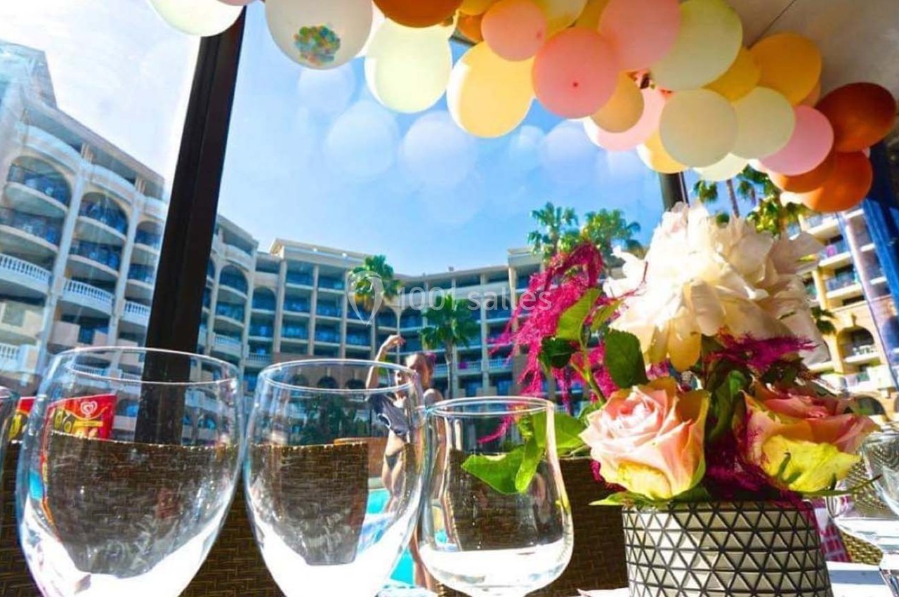 Verres et bouquet de fleurs sur une table, décorée avec des ballons colorés, près d'une piscine et d'un bâtiment.