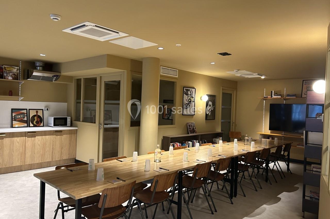 Salle de réunion lumineuse avec une grande table en bois, chaises, étagères, cuisine équipée et décorations murales.