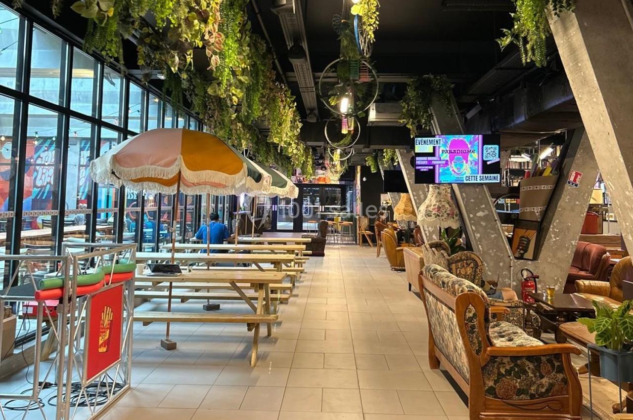 Intérieur d'un espace de restauration avec tables en bois, parasols colorés et décorations végétales suspendues.