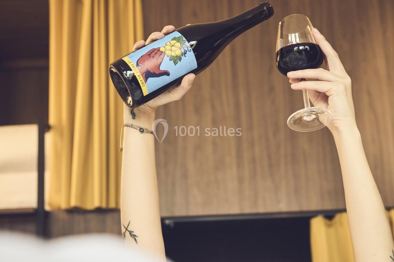 Une personne tient une bouteille de vin et un verre rempli de vin rouge devant un rideau jaune.