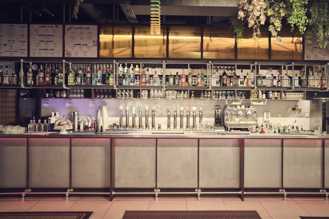 Bar moderne avec comptoir en métal, rangées de bouteilles d'alcool, tireuses à bière et décoration suspendue.