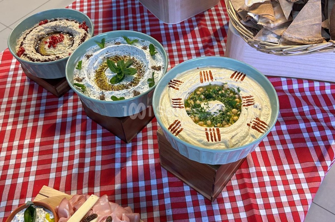 Trois bols de houmous garnis de différentes herbes et épices, présentés sur une nappe à carreaux rouges et blancs.