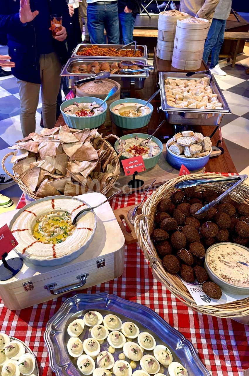 Buffet varié avec houmous, falafels, pains pita, mezzés, œufs farcis et plats chauds sur une nappe à carreaux rouges.
