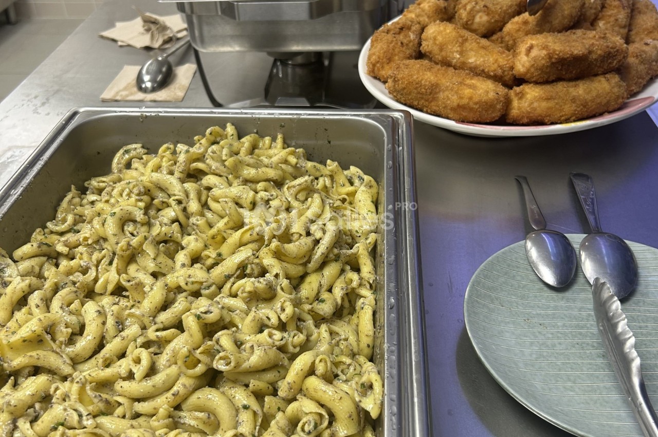 Plat de pâtes assaisonnées aux herbes dans un bac en inox, accompagné d'une assiette de croquettes dorées.