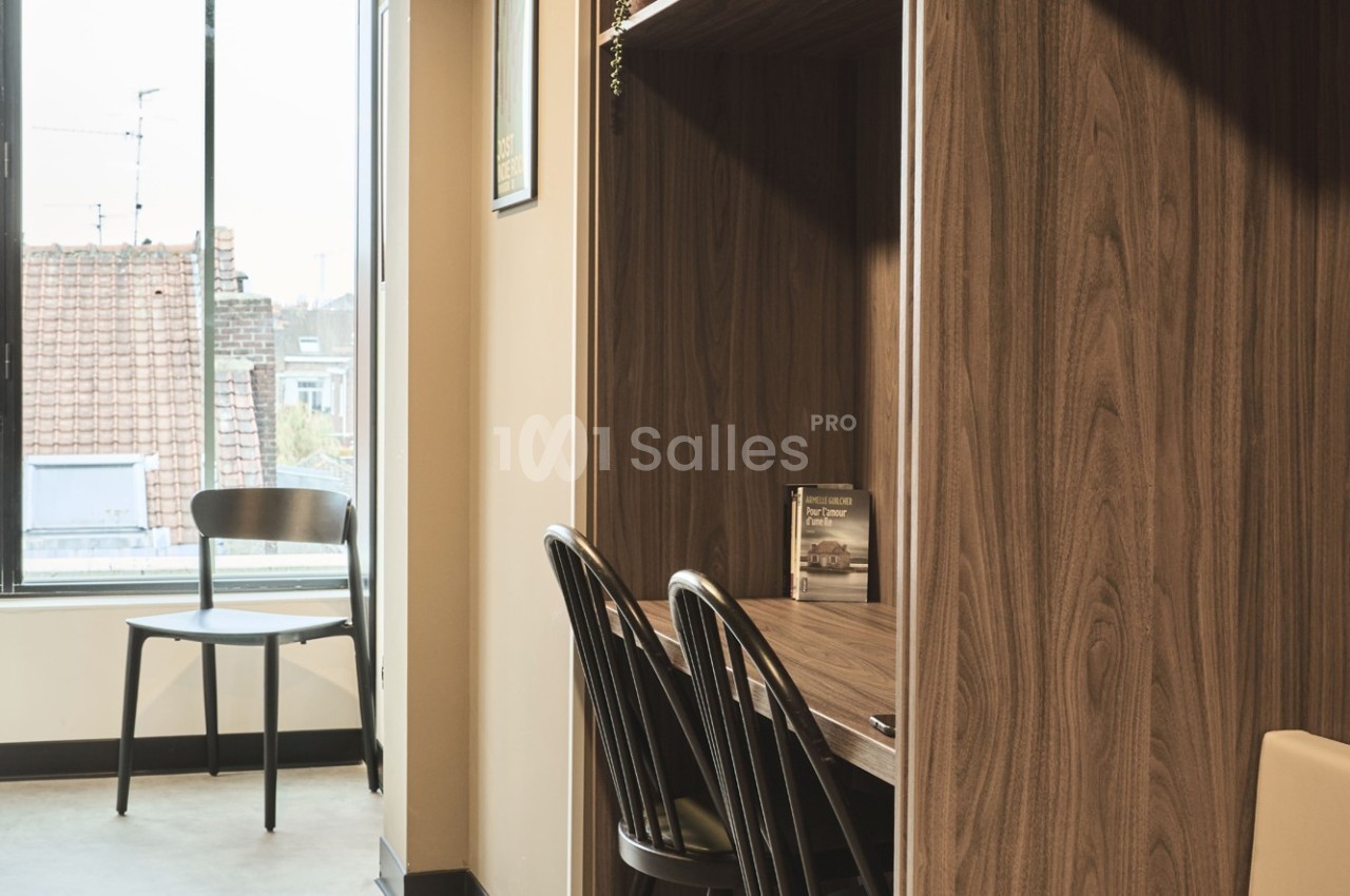 Bureau en bois avec deux chaises noires, près d'une fenêtre offrant une vue sur des toits et un ciel dégagé.