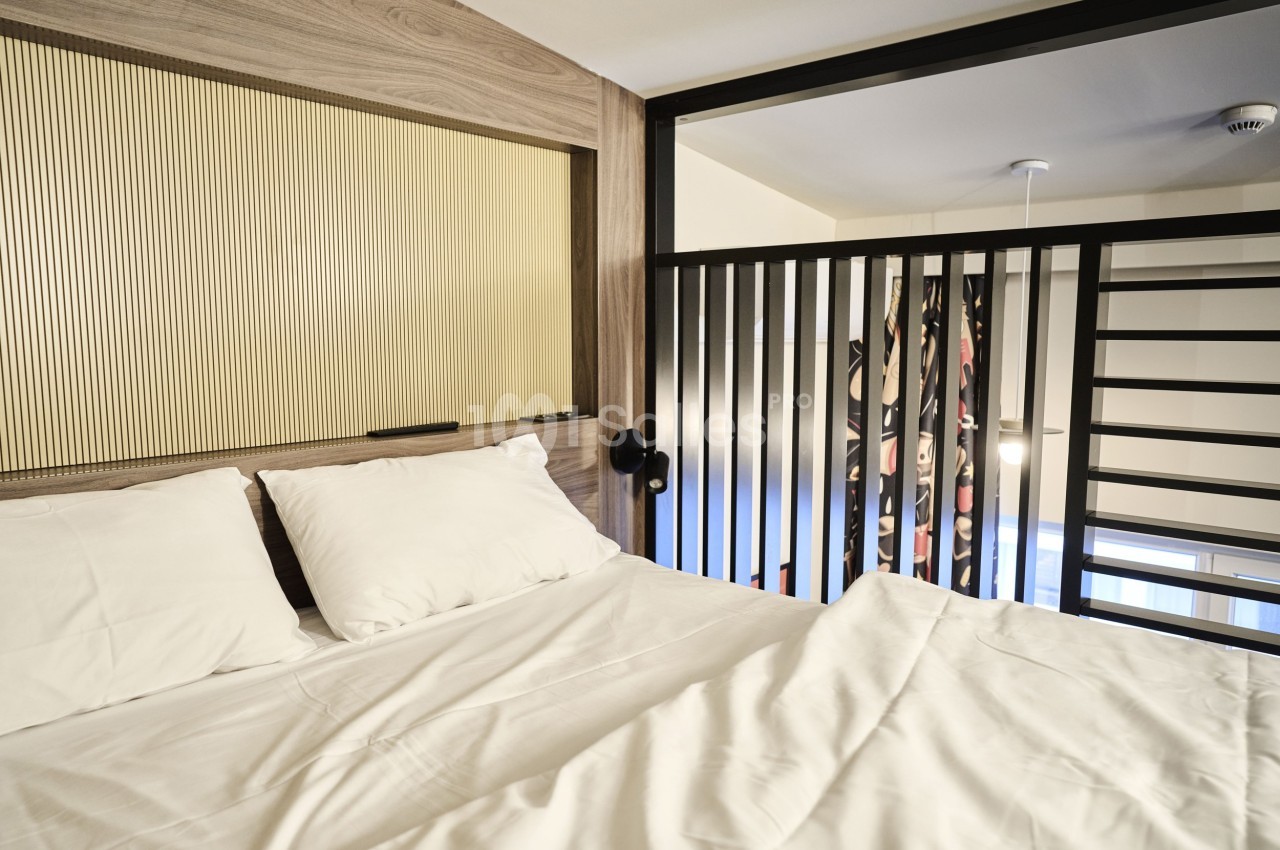 Lit double avec draps blancs dans une chambre moderne, séparée par une cloison en bois et une rambarde métallique.