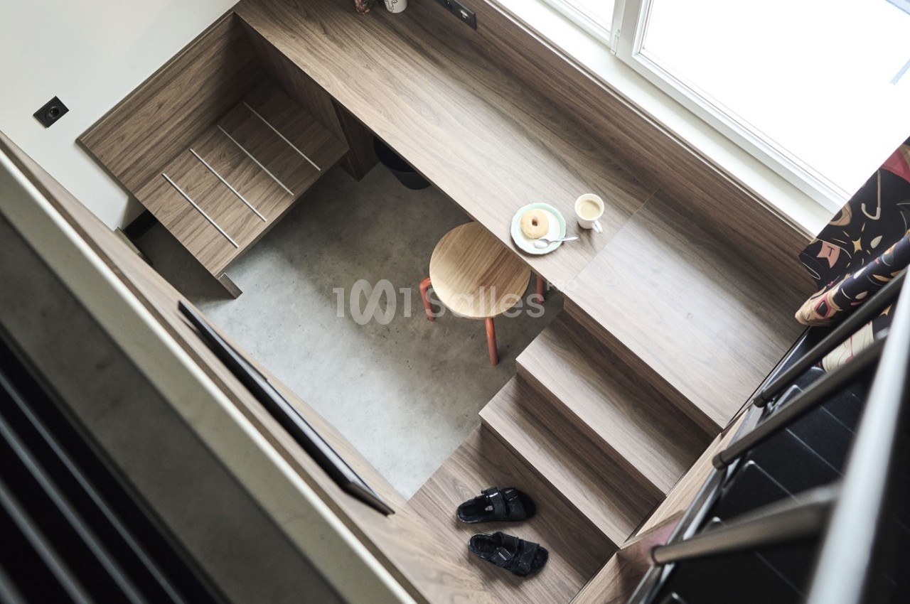 Vue plongeante sur un espace de travail compact avec bureau en bois, tabouret, tasse, assiette et escaliers intégrés.