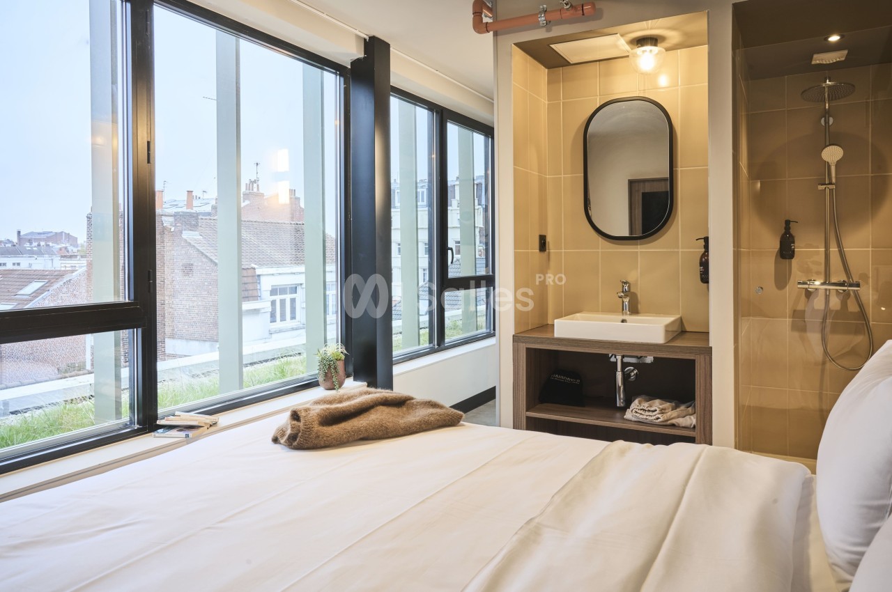 Chambre lumineuse avec lit, lavabo, miroir et douche, donnant sur de grandes fenêtres avec vue sur des toits urbains.