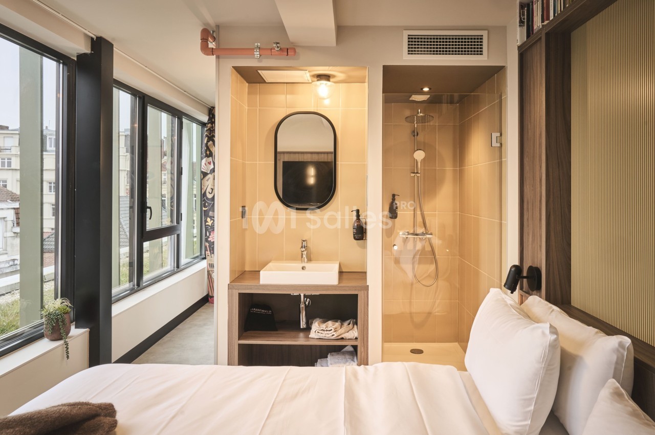 Chambre lumineuse avec lit, salle de bain ouverte comprenant une douche, un lavabo et un miroir, vue sur des fenêtres.