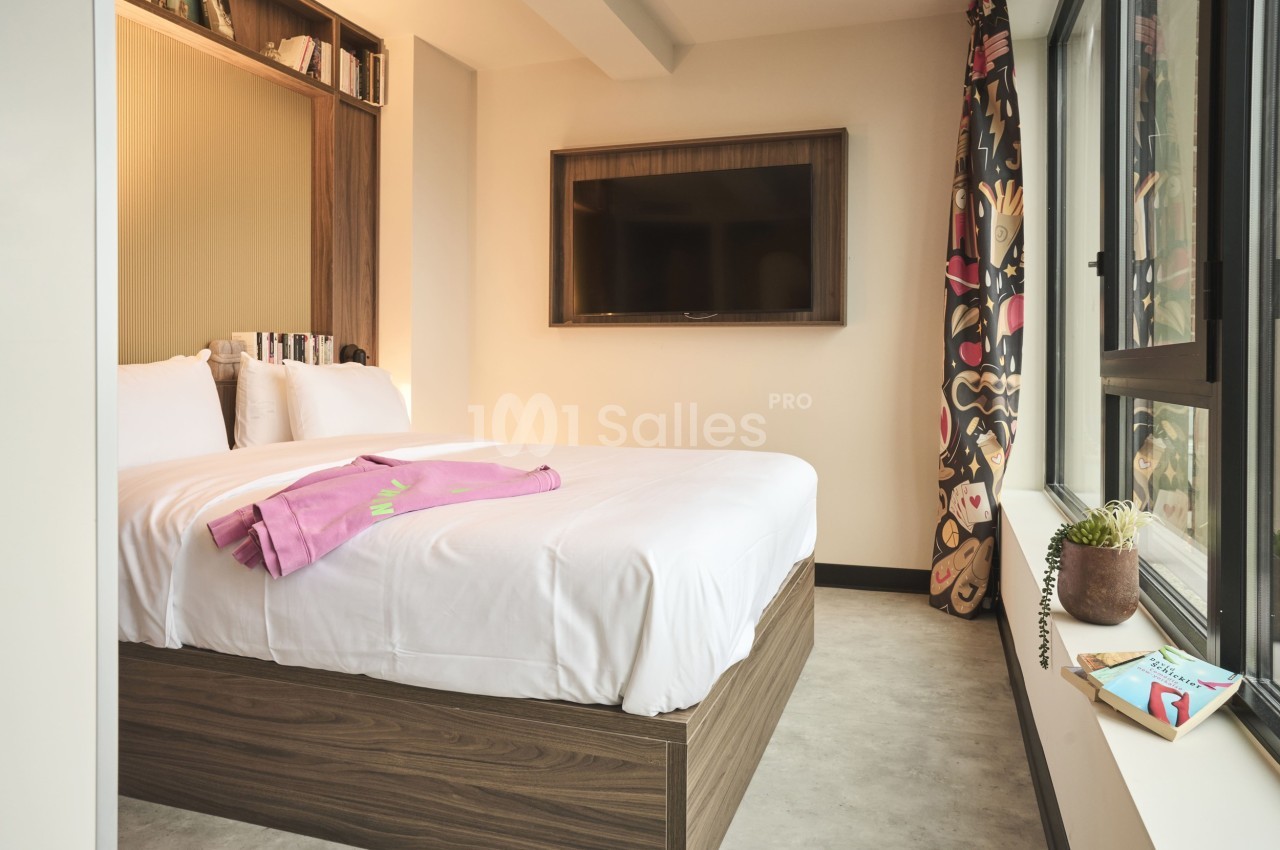 Chambre lumineuse avec lit double, couverture rose, étagères intégrées, télévision murale et grande fenêtre avec rideaux…