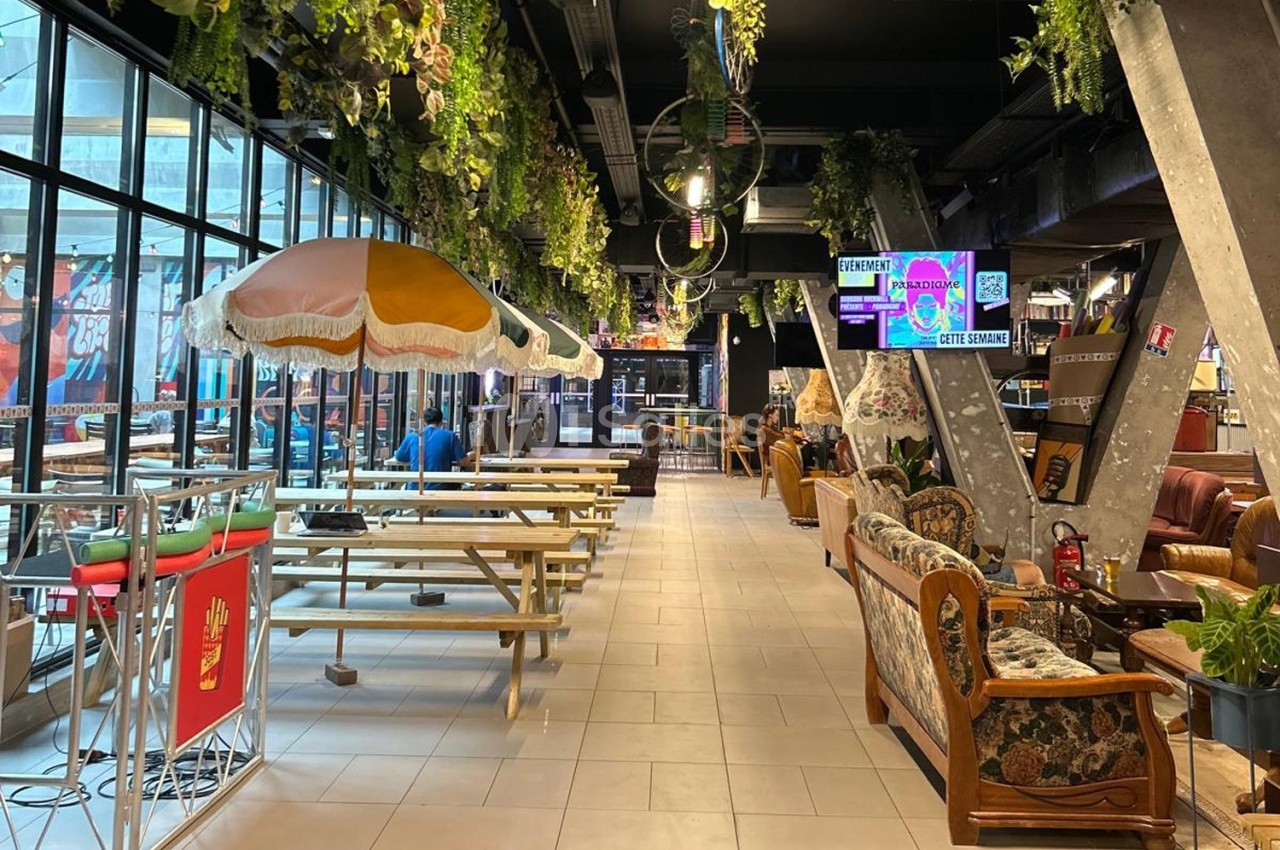 Intérieur d'un espace de restauration avec tables en bois, parasols colorés et décorations végétales suspendues.