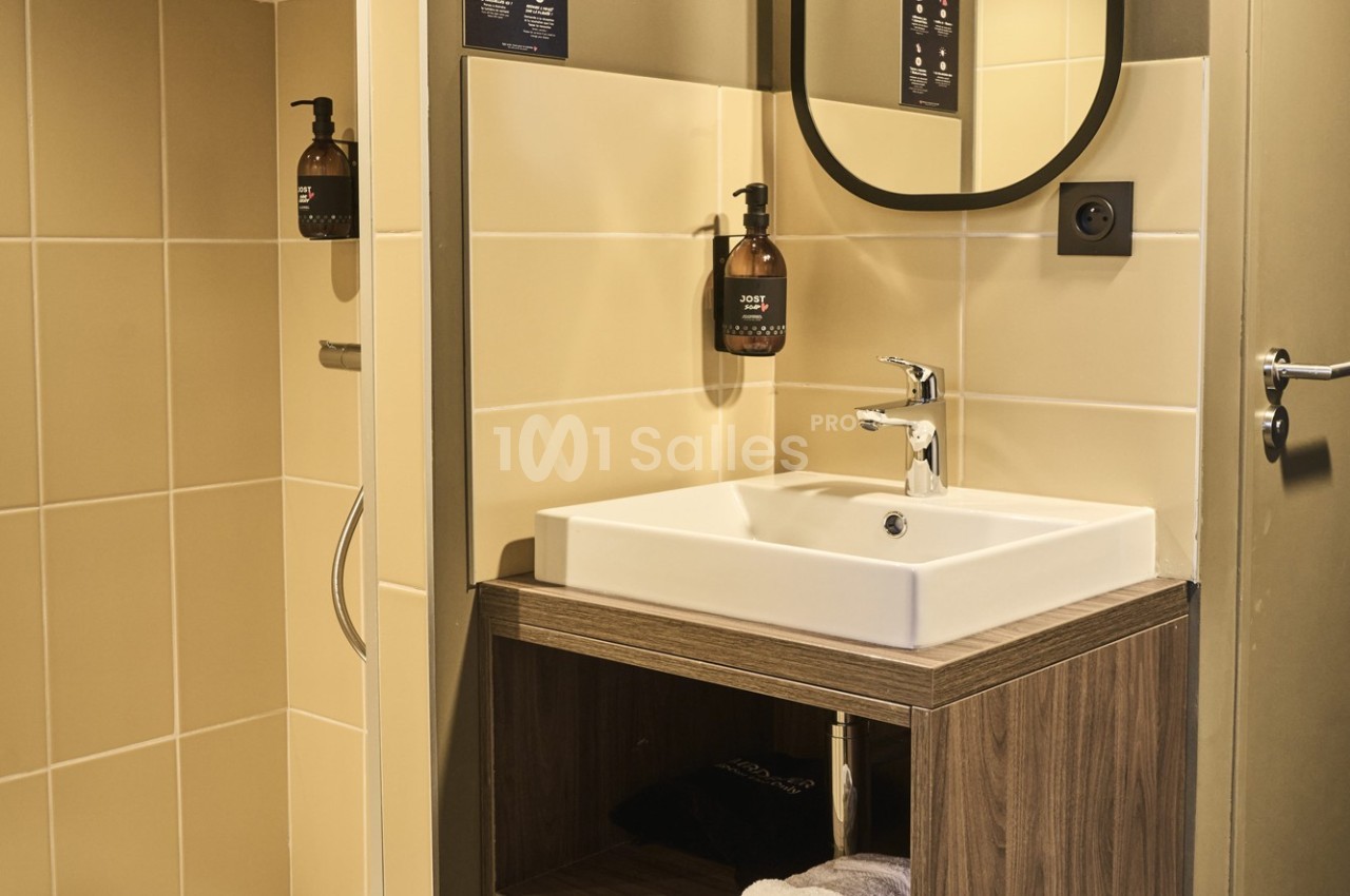 Lavabo moderne avec meuble en bois, miroir ovale, distributeur de savon mural et carrelage beige.