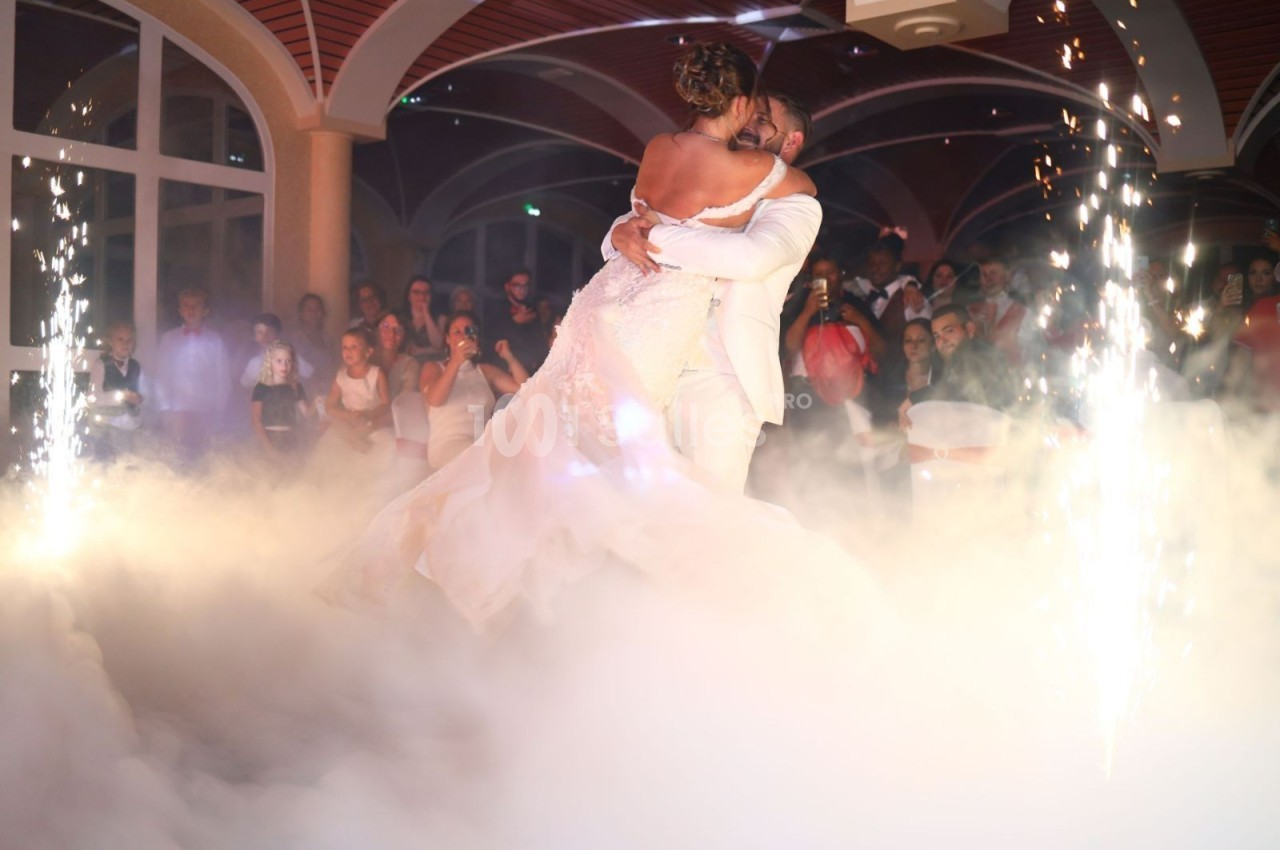 Un couple de mariés danse au milieu de la fumée et des étincelles, entouré d'invités dans une salle festive.