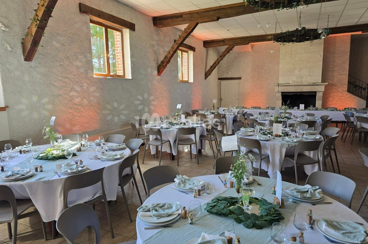 Salle de réception décorée pour un événement, avec tables rondes dressées, nappes blanches et éléments végétaux.