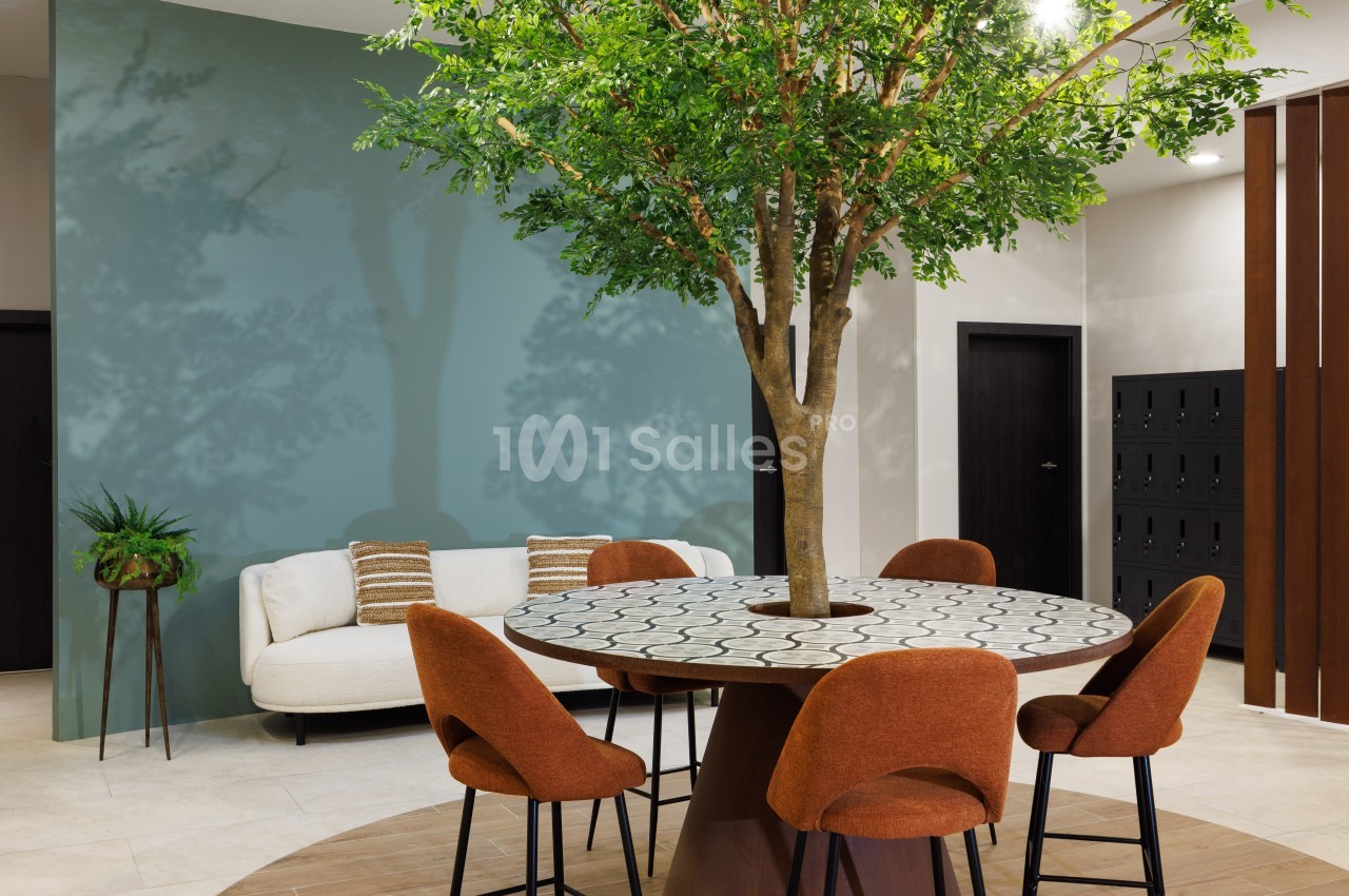 Table ronde avec un arbre en son centre, entourée de chaises orange, dans un espace intérieur moderne avec un mur bleu.