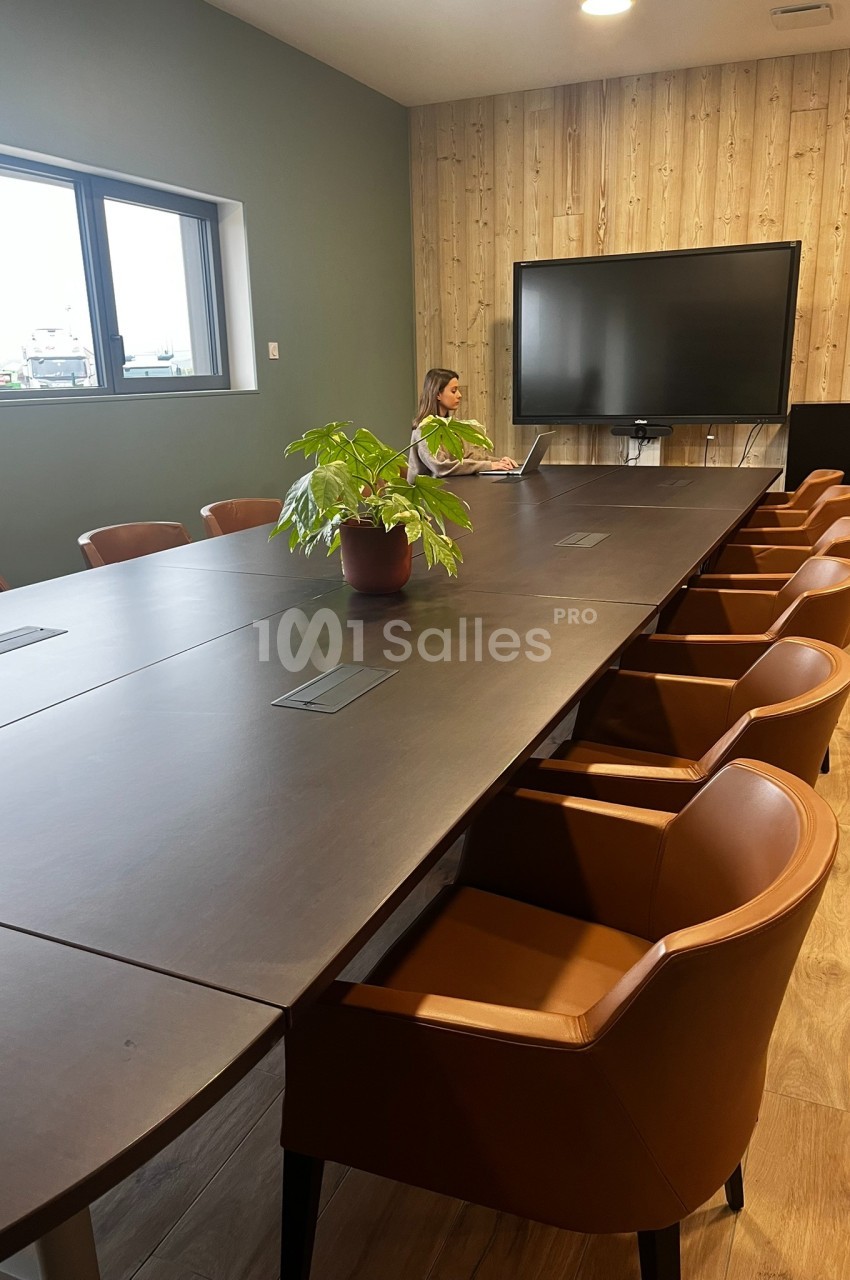 Salle de réunion avec une grande table en bois sombre, des chaises en cuir marron, une plante et un écran au mur.
