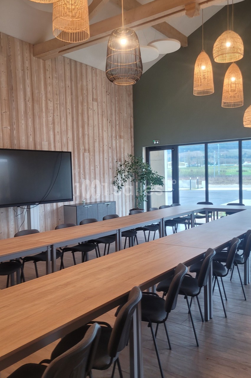 Salle de réunion lumineuse avec de longues tables en bois, chaises noires, luminaires suspendus et grande baie vitrée.